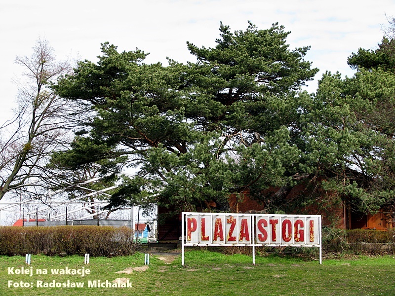 Napis „Plaża Stogi” przy wejściu na teren kąpieliska w Gdańsku, w tle widoczne dojście do plaży i zieleń.