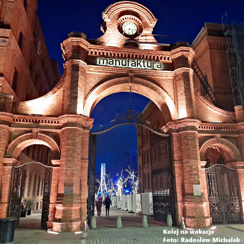 Podświetlone wejście do Centrum Handlowego Manufaktura w Łodzi nocą. Historyczne czerwone mury z cegły dawnej fabryki Poznańskiego są efektownie oświetlone ciepłym światłem, kontrastującym z ciemnym niebem.