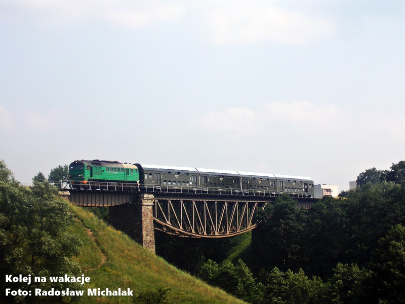 Historyczna fotografia pociągu specjalnego z lipca 2010 roku. Spalinowa lokomotywa ST44-949 z oliwkowymi wagonami piętrowymi Bipa przejeżdża przez wysoki, stalowy most kratownicowy nad rzeką Brdą w okolicy Koronowa. Skład znajduje się wysoko nad lustrem wody, otoczony gęstymi drzewami. Linia ta jest obecnie wyłączona z ruchu.