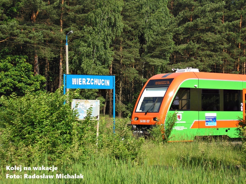 Spokojny kadr ze stacji kolejowej Wierzchucin ukrytej w lesie. Na pierwszym planie widoczny bok szynobusu SA106-117 relacji do Bydgoszczy. Centralny punkt tła stanowi niebieska tablica z nazwą miejscowości, otoczona ścianą drzew Borów Tucholskich.