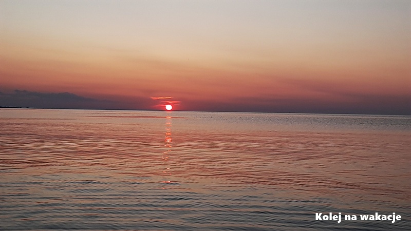 Słońce chowające się za linią horyzontu nad morzem w Jantarze, niebo w odcieniach delikatnego pomarańczu i fioletu.