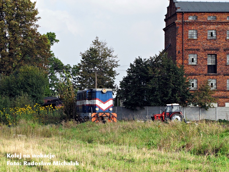 Lokomotywa Lxd2-289 prowadząca pociąg Kaliskiej Kolei Dojazdowej w Goliszewie, na pierwszym planie pole z pracującym traktorem, w tle Młyn Zbożowy.