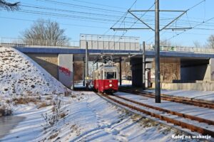 Zabytkowy czerwony tramwaj Konstal 5N z doczepą 5ND przejeżdża pod stalowym wiaduktem kolejowym w Zgierzu. Zimowa sceneria.