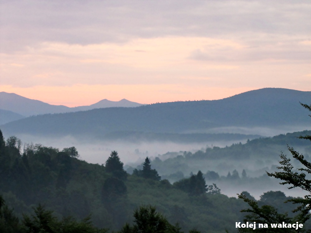 Krajobraz Bieszczadów o świcie w sierpniu. Mgły unoszące się w dolinach między zalesionymi pagórkami pod delikatnym blaskiem wschodzącego słońca, tworzące jesienny klimat w środku lata.