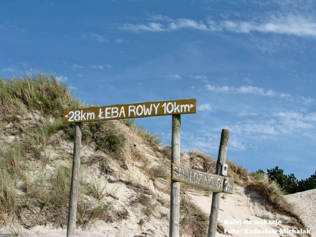 Drewniany drogowskaz na turystycznym szlaku w Słowińskim Parku Narodowym wskazujący odległości: Łeba 28 km, Rowy 10 km, Smołdzino 6 km; w tle piaszczysta wydma i nadmorska roślinność.