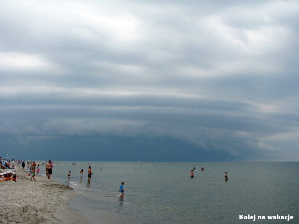 Szeroka, piaszczysta plaża w Jastarni w momencie nadciągającej burzy. Nad ciemnym, wzburzonym Bałtykiem wiszą gęste, granatowe chmury burzowe, kontrastujące z jasnym piaskiem brzegu.