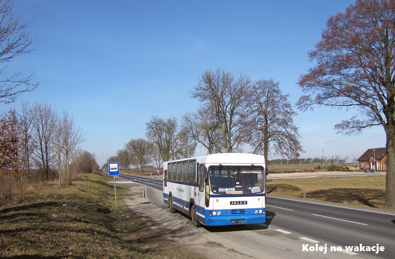 Zdjęcie z marca 2013 roku przedstawiające autobus Jelcz L11 w barwach PKS Piotrków Trybunalski jadący drogą, obok której wyraźnie widać podwyższony nasyp po zlikwidowanym torowisku kolei wąskotorowej.