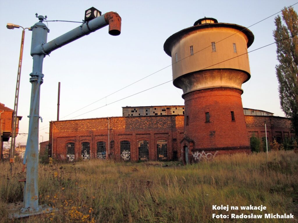 Zabytkowa parowozownia w Gnieźnie o zmierzchu: wieża ciśnień, hala wachlarzowa i żuraw wodny w otoczeniu wysokiej trawy.