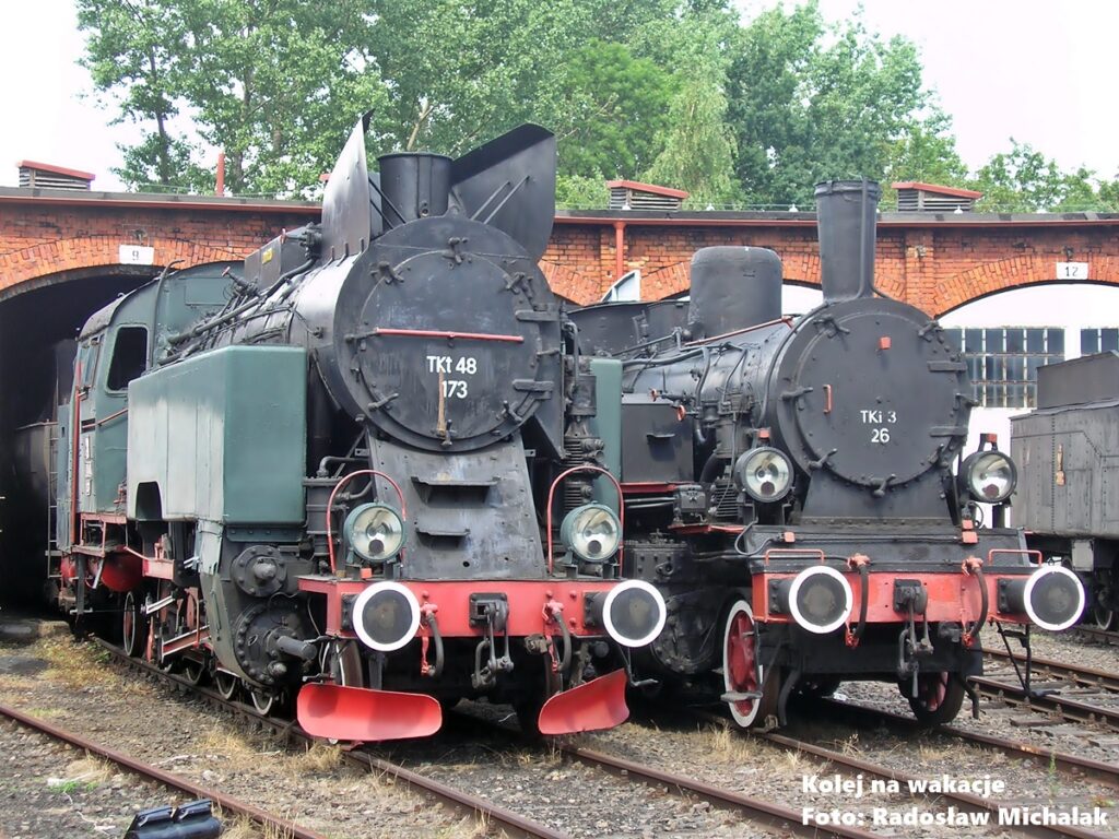 Zabytkowe parowozy Tkt48-173 oraz TKi3-26 przed halą parowozowni w Muzeum Kolejnictwa w Jaworzynie Śląskiej. Eksponaty na torach zewnętrznych skansenu.
