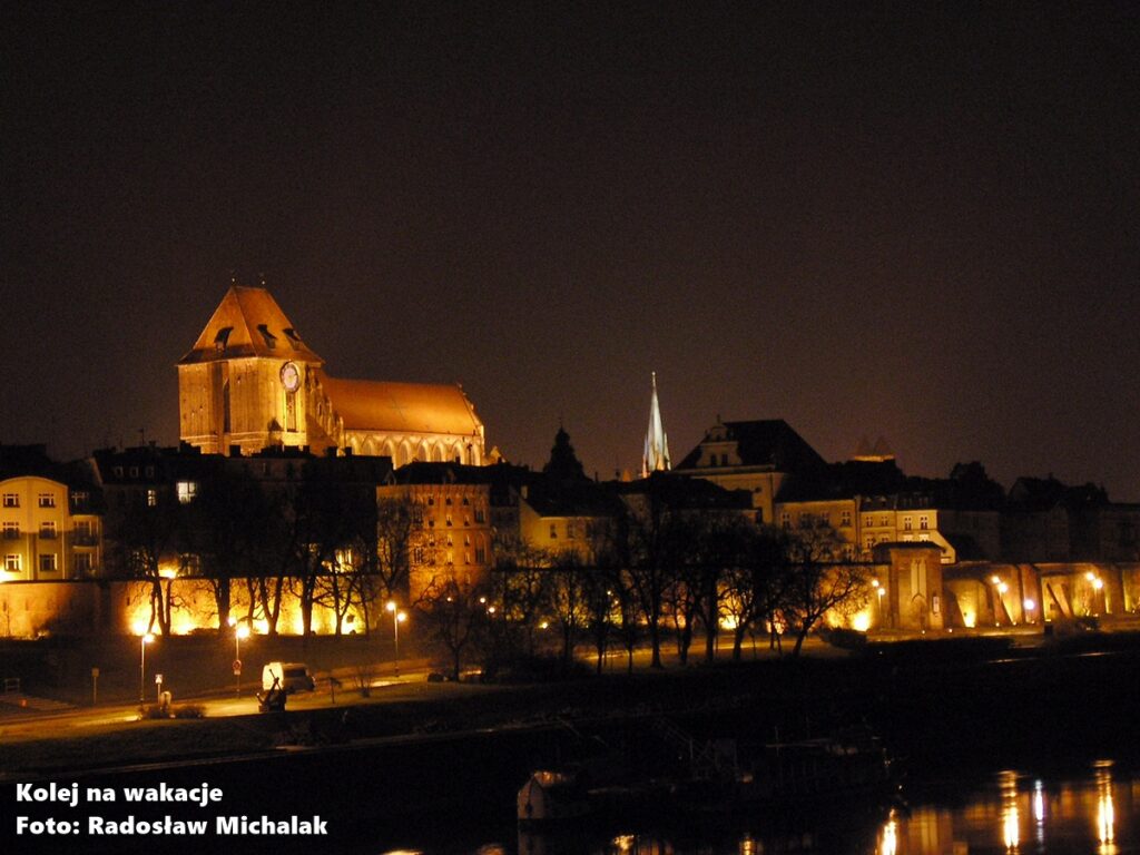 Nocna panorama Starego Miasta w Toruniu widoczna z mostu nad Wisłą, oświetlone zabytki odbijające się w wodzie.