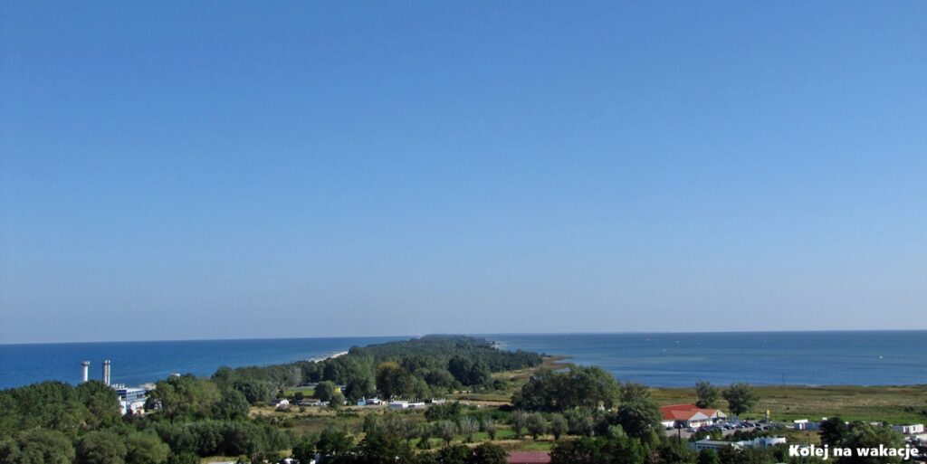 anorama Półwyspu Helskiego widziana z lotu ptaka z wieży we Władysławowie. Wąski pas lądu porośnięty lasem ciągnie się ku horyzontowi, oddzielając otwarte, wzburzone Morze Bałtyckie po lewej stronie od spokojnych wód Zatoki Puckiej po prawej.