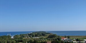 anorama Półwyspu Helskiego widziana z lotu ptaka z wieży we Władysławowie. Wąski pas lądu porośnięty lasem ciągnie się ku horyzontowi, oddzielając otwarte, wzburzone Morze Bałtyckie po lewej stronie od spokojnych wód Zatoki Puckiej po prawej.