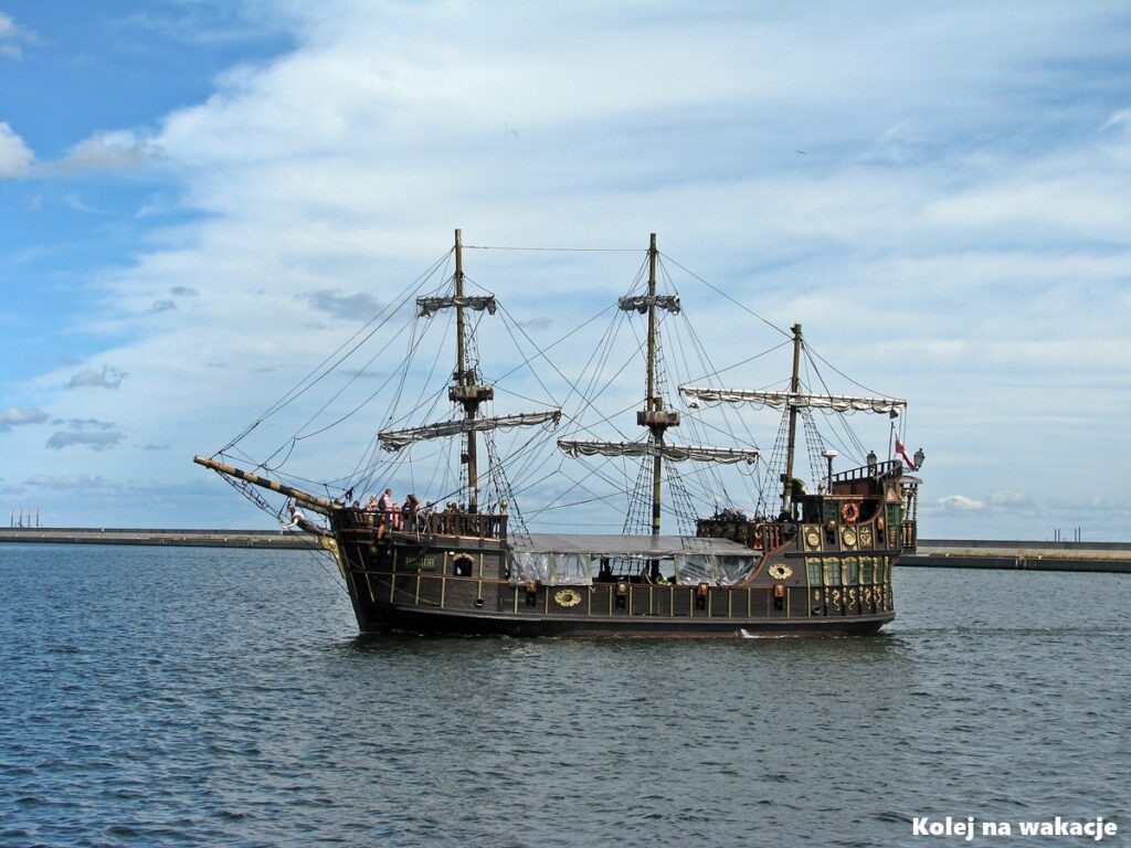 Drewniany statek turystyczny stylizowany na galeon wpływający do portu w Gdyni, mijający falochron na tle błękitnego nieba.
