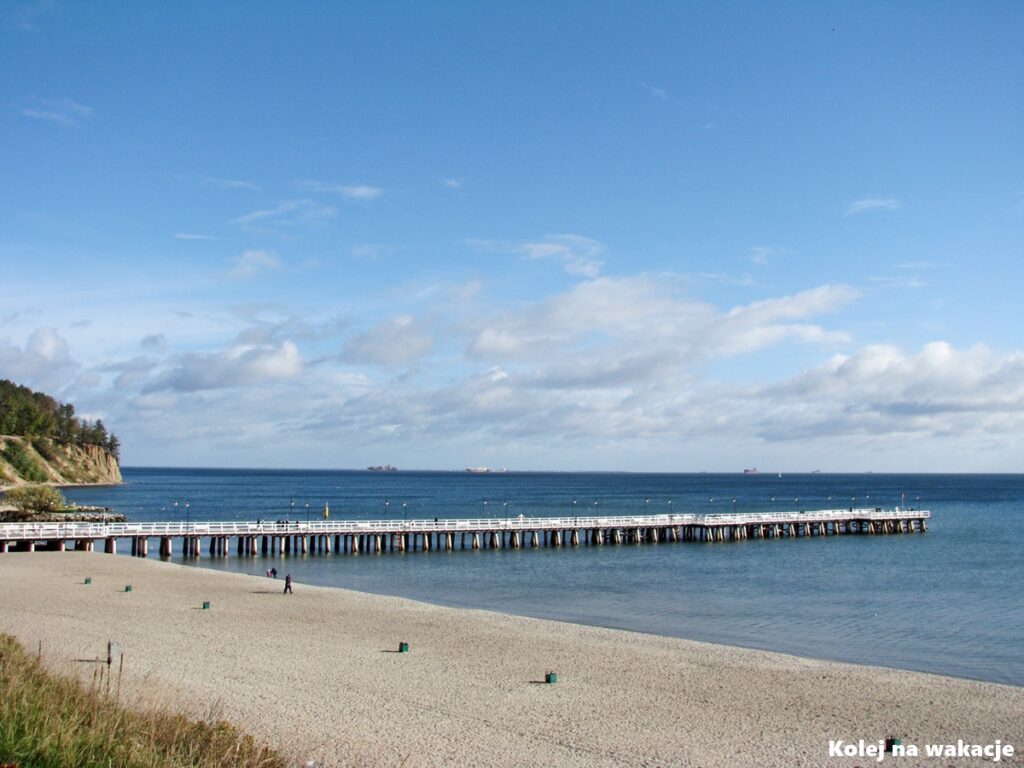 Drewniane molo spacerowe w gdyńskiej dzielnicy Orłowo, po lewej stronie piaszczysta plaża, w tle zalesiony, stromy brzeg klifu orłowskiego.