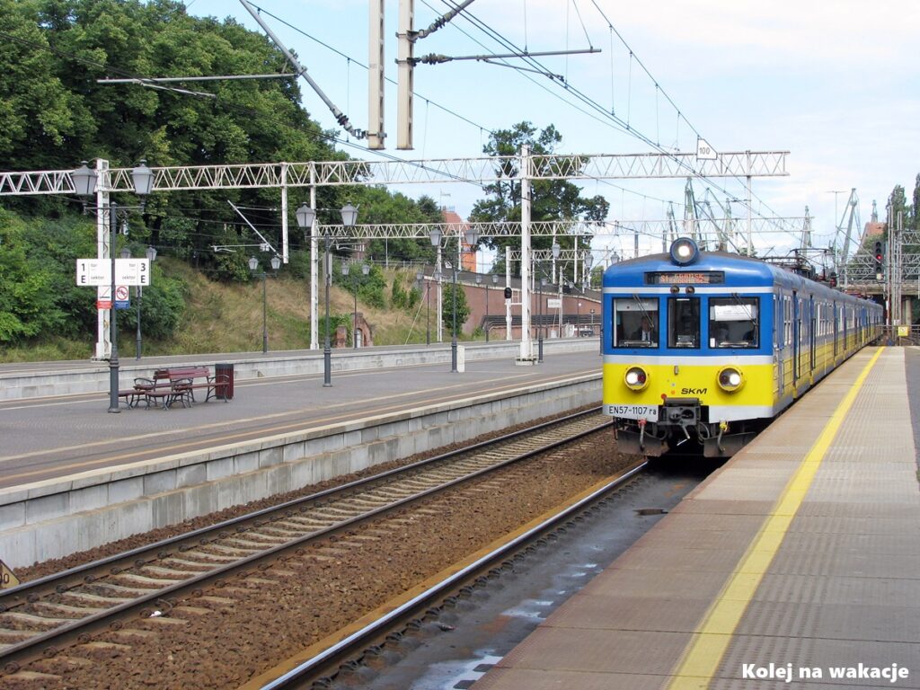 Klasyczny elektryczny zespół trakcyjny EN57-1107 z dużymi reflektorami (tzw. wiadrami) w tradycyjnym, żółto-niebieskim malowaniu SKM Trójmiasto. W tle widoczna infrastruktura i żurawie stoczniowe.