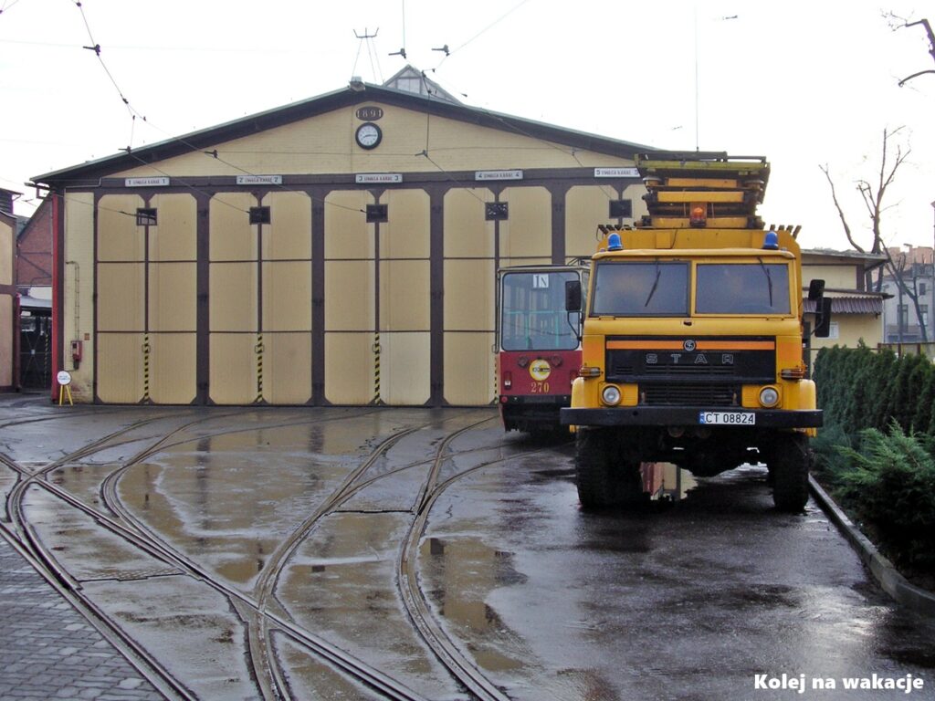 Ceglana hala zajezdni tramwajowej w Toruniu, przed którą stoi techniczny samochód ciężarowy marki Star z podnośnikiem do naprawy sieci trakcyjnej.