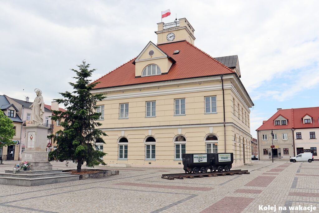 Rynek w Łęczycy z klasycystycznym ratuszem oraz wagonikami górniczymi przypominającymi o przemysłowym dziedzictwie regionu.